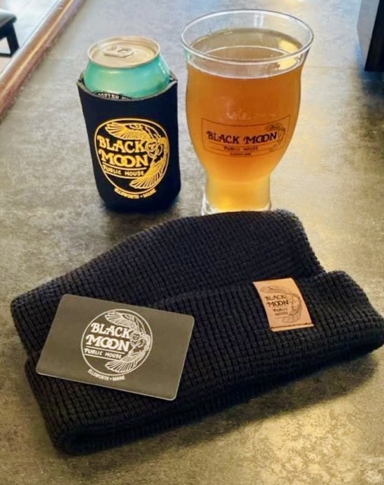 A navy waffle-knit beanie with a leather logo tab, beside a koozie and a beer