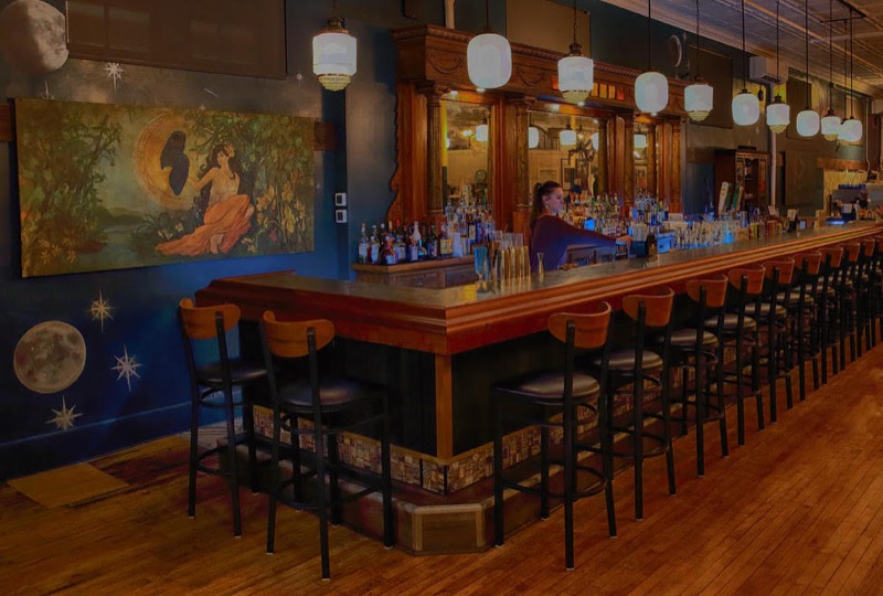 The long wooden bar under milk-glass pendant lights, with a mural and a teal wall painted with moons and stars