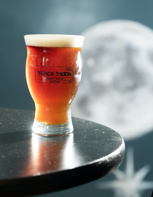 A pint of red ale in a Black Moon glass on a black table, in front of a painted full moon