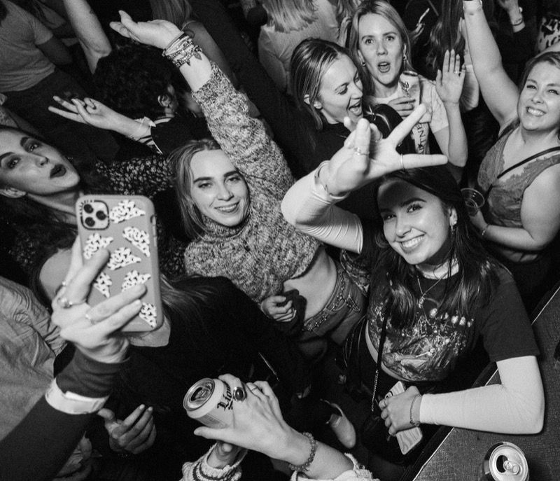 Black and white photo of a crowd of women dancing and cheering, one taking a selfie