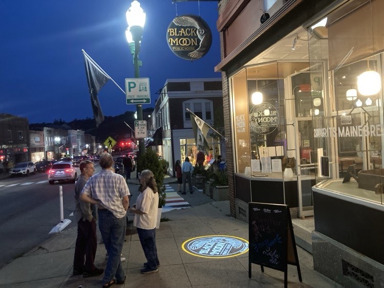 The Black Moon storefront on Main Street at night, with people on the sidewalk and the logo projected onto the pavement
