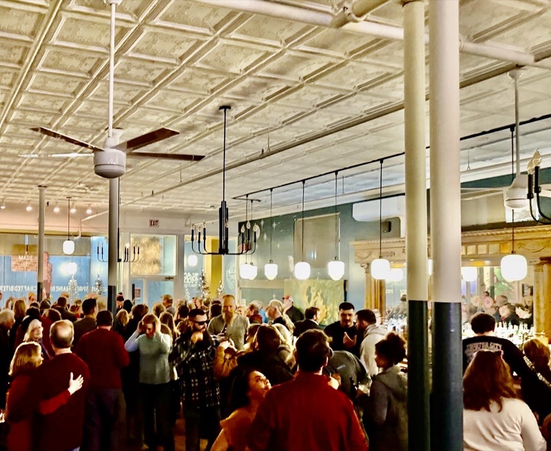 A packed room of people standing under the pressed tin ceiling