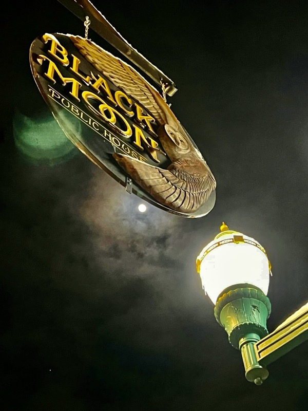 The round Black Moon sign hanging over Main Street at night, with a street lamp and the moon behind clouds